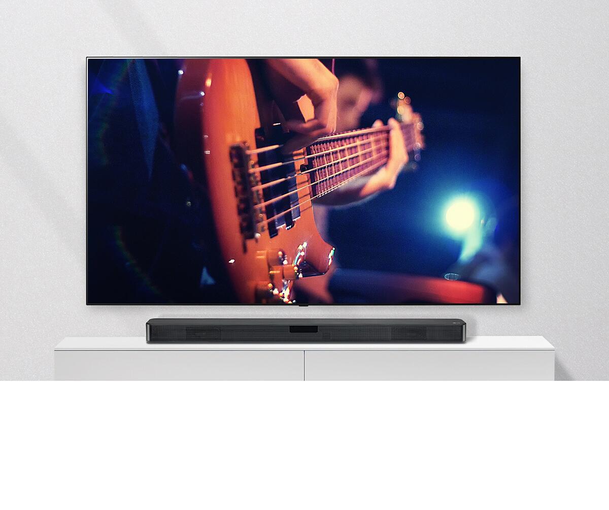 The TV is attached to the wall, and the sound bar is on a white shelf. TV showing a man plays guitar.