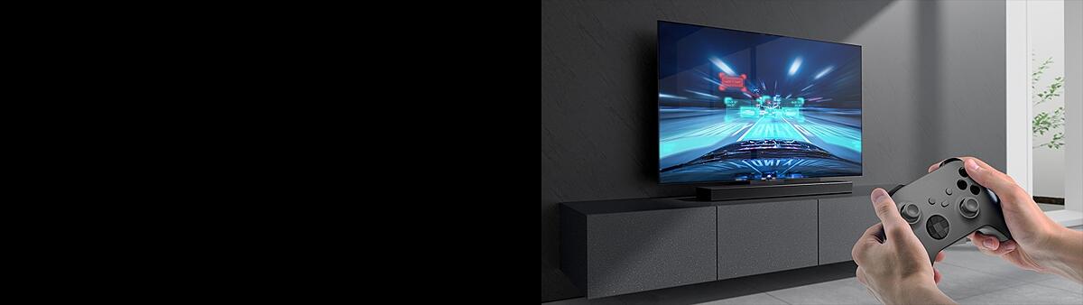 The LG Soundbar is placed on the cabinet and racing game scene is shown on the LG TV coneected to the Soundbar . A game console is on the bottom right side of the picture hold by two hands.