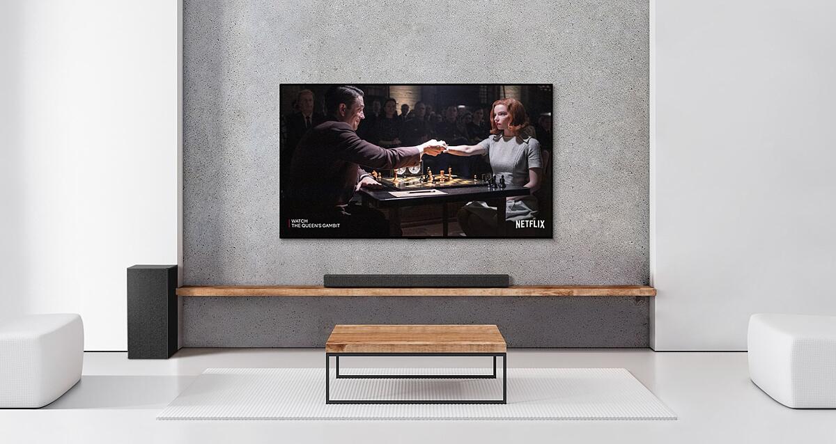 A set of 2 rear speakers, subwoofer, and a soundbar, and TV are in a white living room. A poster of a TV show is on TV screen.