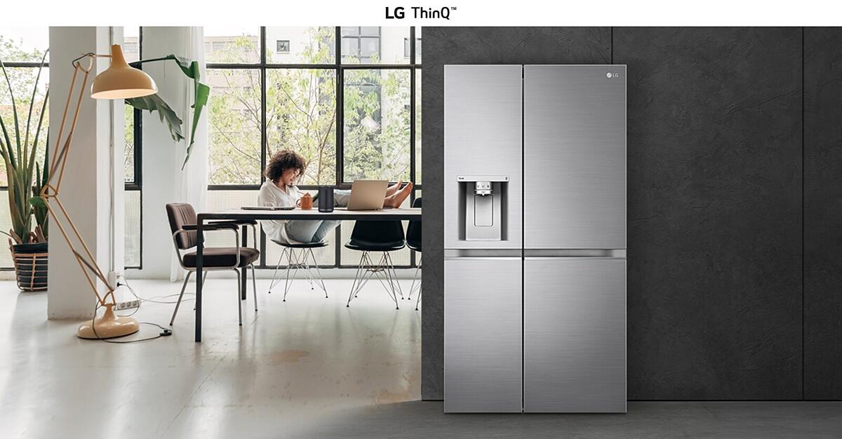 Stainless steel LG ThinQ fridge beside a large window with greenery, person working on a laptop at a modern table.