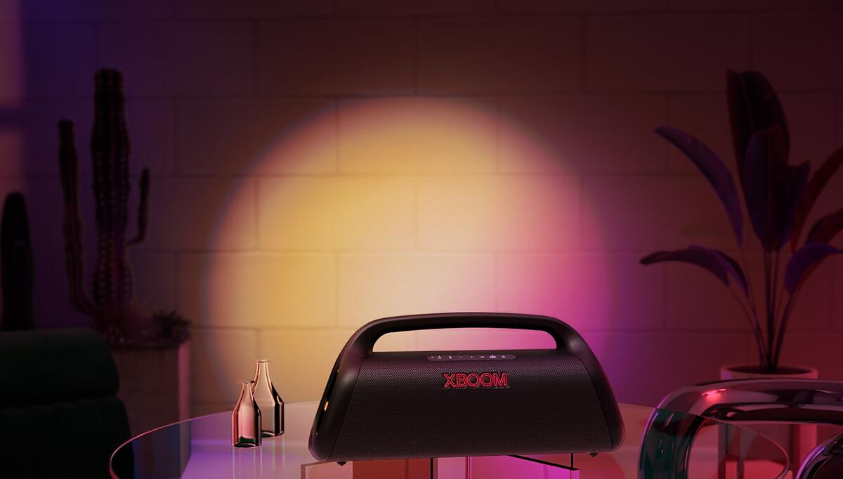 An image of LG XBOOM Go XG9. It's placed on the glass table at home, casts orange and pink gradient stage lighting.